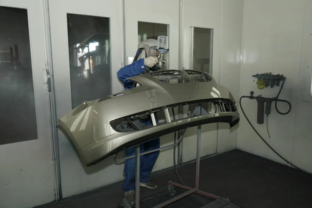A person wearing protective gear uses a spray gun to paint a car bumper in an industrial paint booth.