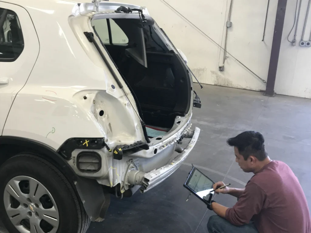 A person crouches next to a white SUV with its rear bumper and hatch removed, inspecting or taking notes on a clipboard in a garage or repair shop.