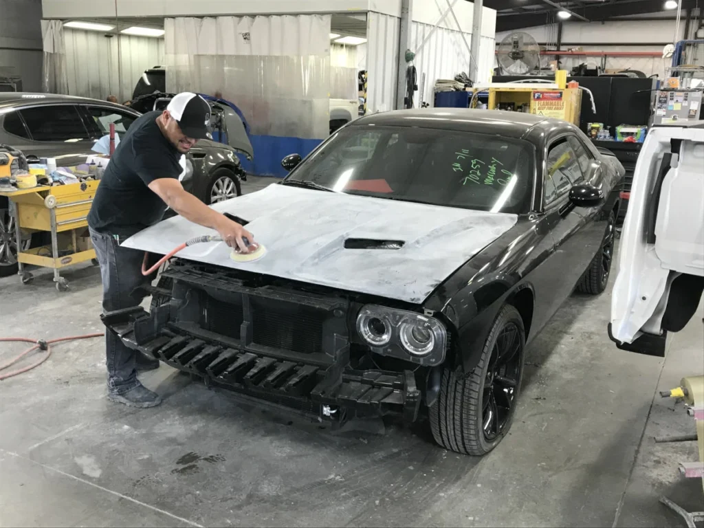 A person sands the hood of a black car with a power tool in an auto body shop. The cars front bumper is removed, and various equipment and other vehicles are visible in the background.