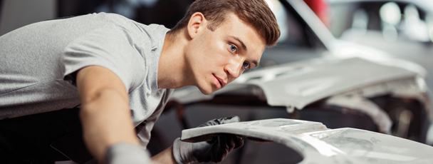 A young man wearing gloves carefully inspects and works on a car body part in an auto repair shop, focusing intently on the task.