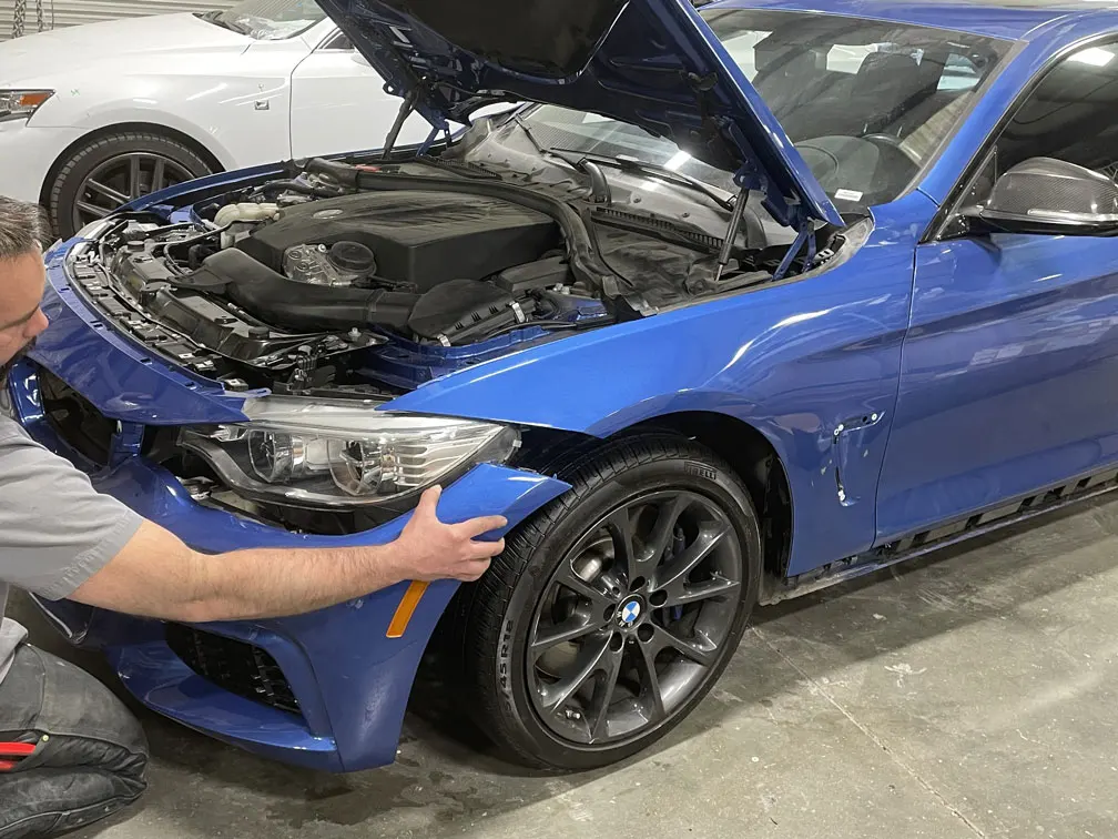 A person inspects the damaged front bumper and fender of a blue car with its hood open inside a garage. Another white car is parked nearby in the background.